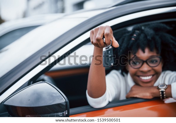 Young african american woman sits inside of new\
modern car with keys in\
hand.