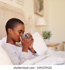 Young African American woman sitting up in her bed in the morning sipping a cup of coffee - Powered by Shutterstock