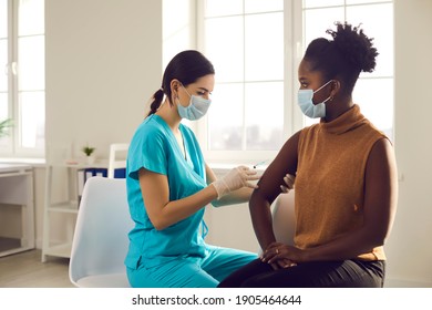 Young African American woman sitting at doctor's office and getting a flu shot in her arm. Nurse in medical face mask holding syringe and giving female patient modern Covid 19 vaccine injection