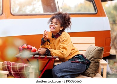 Young African American Woman Sitting At Table Near Car In Autumn Forest And Drinking Hot Tea, Dreamy Mixed Race Female Relaxing Around Campsite, Spending Time In Nature. Camping, Travel And Vanlife