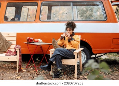 Young african american woman sitting at table near car in autumn forest and drinking hot tea, dreamy female relaxing around campsite, spending time in nature. Camping, travel and vanlife - Powered by Shutterstock