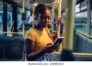 Young african american woman smiling while standing by herself on a bus and using a smartphone to send a message. Black female passenger browsing social media in public transportation during a night. - Powered by Shutterstock