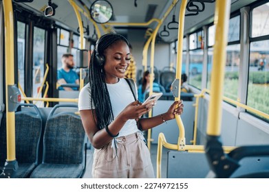 Young african american woman smiling while standing by herself on a bus and listening to music on a smartphone. Happy female passenger listening to music on a smartphone in public transportation. - Powered by Shutterstock