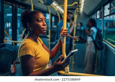 Young african american woman smiling while standing by herself on a bus and listening to music on a smartphone in the night. Female passenger using airpods and mobile phone in public transportation. - Powered by Shutterstock