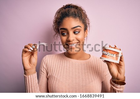 Young african american woman smiling happy holding professional orthodontic denture with metal braces and removable invisible aligner. Comparation of two dental straighten treatments