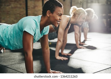 Young African American woman in sportswear smiling while doing pushups with a group of friends during a workout class at the gym - Powered by Shutterstock