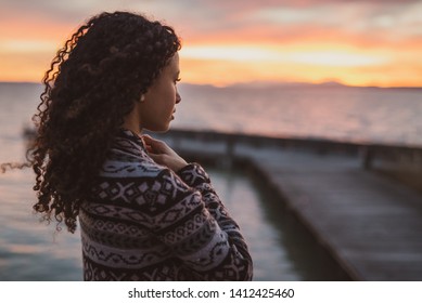 Young African American woman is standing on the promenade at the lake, looking thoughtfully towards the water and the setting sun. The girl in a vest is in a thoughtful mood - Shutterstock ID 1412425460
