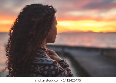 Young African American Woman Is Standing On The Promenade At The Lake, Looking Thoughtfully Towards The Water And The Setting Sun. The Girl In A Vest Is In A Thoughtful Mood