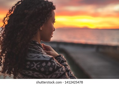 Young African American woman is standing on the promenade at the lake, looking thoughtfully towards the water and the setting sun. The girl in a vest is in a thoughtful mood