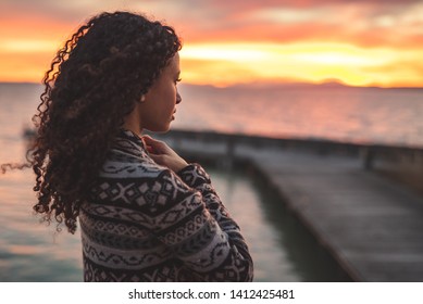 Young African American woman is standing on the promenade at the lake, looking thoughtfully towards the water and the setting sun. The girl in a vest is in a thoughtful mood - Shutterstock ID 1412425481