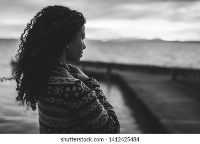 Young African American Woman Is Standing On The Promenade At The Lake, Looking Thoughtfully Towards The Water And The Setting Sun. The Photo In Black And White