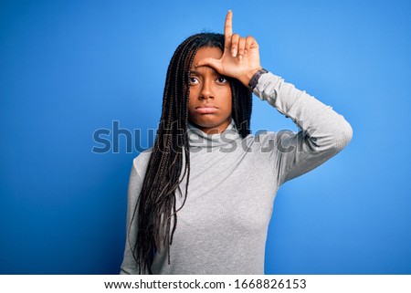 Young african american woman standing wearing casual turtleneck over blue isolated background making fun of people with fingers on forehead doing loser gesture mocking and insulting.