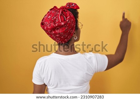 Young african american woman standing over yellow background posing backwards pointing ahead with finger hand 