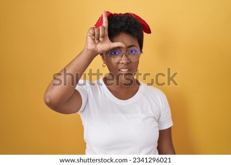 Young african american woman standing over yellow background making fun of people with fingers on forehead doing loser gesture mocking and insulting. 