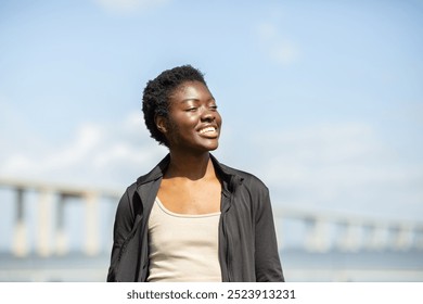 A young African American woman stands by the water under a clear blue sky, smiling joyfully. Behind her, a long bridge stretches across the horizon, creating a picturesque view on a beautiful day. - Powered by Shutterstock