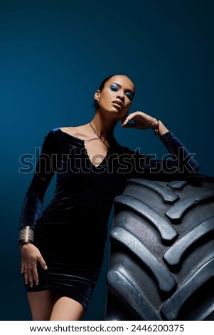 A young African American woman stands confidently next to a colossal tire in a studio setting.