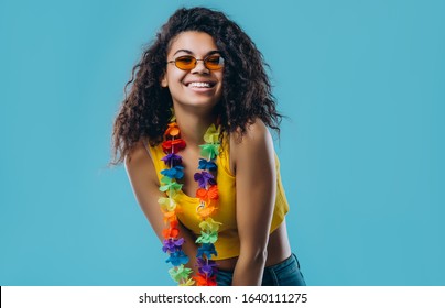 Young African American Woman In Sunglasses With Afro Hair Wearing Flower Hawaiian Lei  Very Happy And Excited Wearing Yellow Top Isolated On Blue Background.