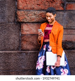 A Young African American Woman is texting on a cell phone outside, wearing an orange, red jacket, a flower-patterned skirt, carrying a laptop computer, standing by a stone wall on campus, smiling.
