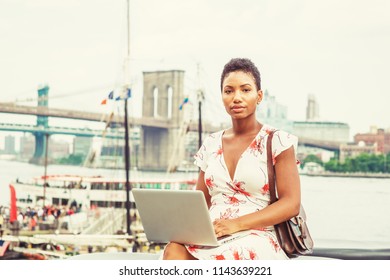 Young African American Woman traveling in New York, with short afro hair, wearing dress, carrying bag, sitting by river working on laptop computer. Brooklyn, Manhattan bridges, boat on background. - Shutterstock ID 1143639221