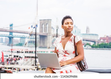Young African American Woman traveling in New York, with short afro hair, wearing dress, carrying bag, sitting by river working on laptop computer. Brooklyn, Manhattan bridges, boat on background.
 - Shutterstock ID 1143639686