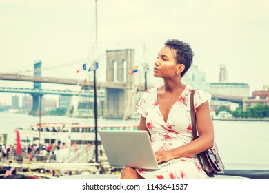 Young African American Woman traveling in New York, with short afro hair, wearing dress, carrying bag, sitting by river working on laptop computer. Brooklyn, Manhattan bridges, boat on background.
 - Shutterstock ID 1143641537