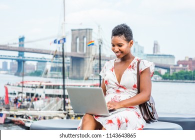 Young African American Woman traveling in New York, with short afro hair, wearing dress, carrying bag, sitting by river, working on laptop computer. Brooklyn, Manhattan bridges, boat on background.
 - Shutterstock ID 1143643865