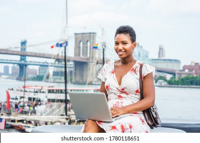 Young African American Woman traveling in New York, with short afro hair, wearing dress, carrying bag, sitting by river, working on laptop computer. Brooklyn, Manhattan bridges, boat on background. - Shutterstock ID 1780118651