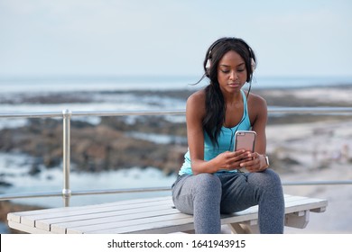 Young african american woman using smartphone on beach female jogger listening to music wearing headphones sitting on bench relaxing by the sea - Powered by Shutterstock