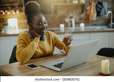 Young African American woman using laptop and toasting with Champagne while talking to someone online. 