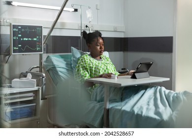 Young african american woman using tablet in hospital ward bed. Sick patient with online technology recovering from disease at clinic with medical equipment. Person web surfing - Powered by Shutterstock