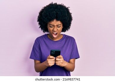 Young African American Woman Using Smartphone Typing Message Angry And Mad Screaming Frustrated And Furious, Shouting With Anger. Rage And Aggressive Concept. 