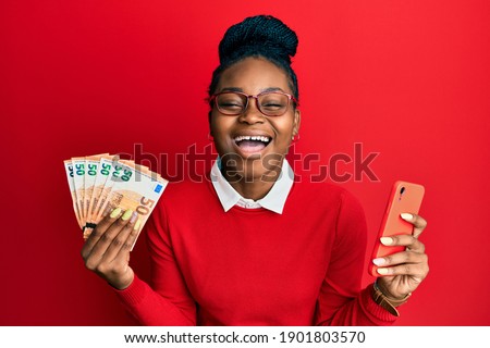Young african american woman using smartphone holding euro banknotes smiling and laughing hard out loud because funny crazy joke. 