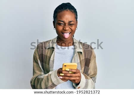 Young african american woman using smartphone sticking tongue out happy with funny expression. 