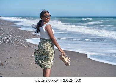Young African American woman walking along the shore of the beach with sandals in hand