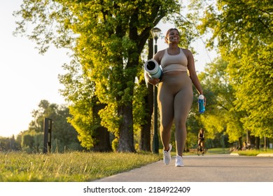 Young African American Woman Walking With Exercise Mat And Bottle Of Water Through The Park In A Summer Day