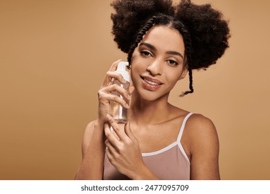 A young African American woman with a warm smile holds a bottle of skincare product against a beige background. - Powered by Shutterstock