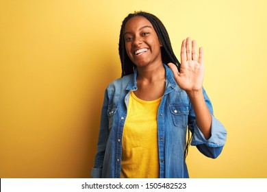 Young african american woman wearing denim shirt standing over isolated yellow background Waiving saying hello happy and smiling, friendly welcome gesture