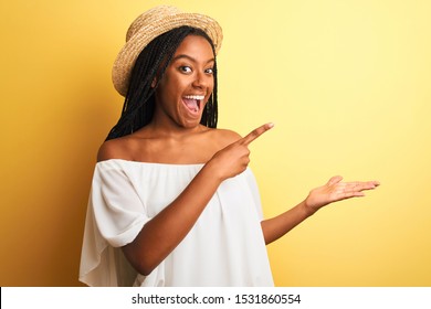 Young african american woman wearing white t-shirt and hat over isolated yellow background amazed and smiling to the camera while presenting with hand and pointing with finger. - Shutterstock ID 1531860554