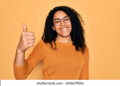 Young african american woman wearing casual sweater and glasses over yellow background doing happy thumbs up gesture with hand. Approving expression looking at the camera showing success.