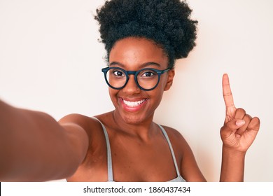 Young african american woman wearing casual clothes and glasses making selfie by the camera surprised with an idea or question pointing finger with happy face, number one 
