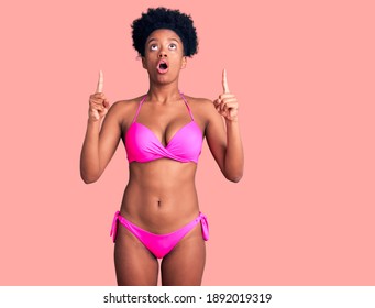 Young African American Woman Wearing Bikini Amazed And Surprised Looking Up And Pointing With Fingers And Raised Arms. 