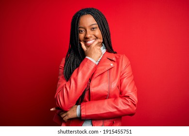 Young african american woman wearing cool fashion leather jacket over red isolated background looking confident at the camera smiling with crossed arms and hand raised on chin. Thinking positive. - Shutterstock ID 1915455751