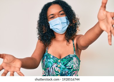 Young African American Woman Wearing Medical Mask Looking At The Camera Smiling With Open Arms For Hug. Cheerful Expression Embracing Happiness. 