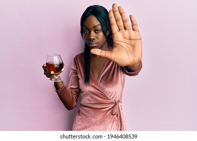 Young african american woman wearing sexy party dress holding cocktail with open hand doing stop sign with serious and confident expression, defense gesture 