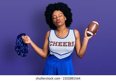 Young african american woman wearing cheerleader uniform holding pompom and football ball looking at the camera blowing a kiss being lovely and sexy. love expression. 