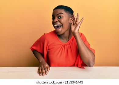 Young African American Woman Wearing Casual Clothes Sitting On The Table Smiling With Hand Over Ear Listening And Hearing To Rumor Or Gossip. Deafness Concept. 