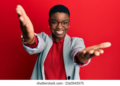 Young african american woman wearing business jacket and glasses looking at the camera smiling with open arms for hug. cheerful expression embracing happiness. 