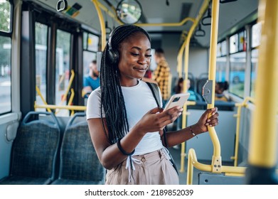 Young African american woman wearing earphones and listening to music on a smartphone while standing alone on a bus. Traveling to work and enjoying a bus ride. Portrait of a beautiful black woman. - Powered by Shutterstock