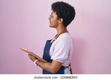 Young african american woman wearing professional waitress apron holding clipboard looking to side, relax profile pose with natural face and confident smile. 