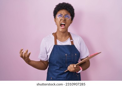 Young african american woman wearing professional waitress apron holding clipboard angry and mad screaming frustrated and furious, shouting with anger. rage and aggressive concept.  - Shutterstock ID 2310907285